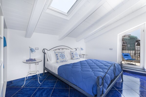 Quarto com claraboia, cama com roupa de cama azul, uma pequena mesa de cabeceira e vista para a varanda.