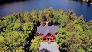 Un chalet avec un bâtiment principal entouré d’une forêt dense, un lac en arrière-plan et une petite cabane sur la gauche.