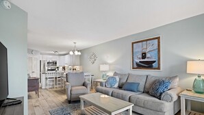 A living room with a grey sofa, a coffee table, and a TV. There is a kitchen in the background with white cabinets and a dining area with a white table and chairs. - Beautiful Spacious Condo Overlooking Pool - walk to Beach and  Coligny Plaza ! (Hilton Head Island)