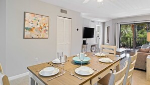 Uma sala de jantar com uma mesa de madeira posta para quatro pessoas, um ventilador de teto, uma televisão e uma varanda com área de estar externa.