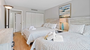 Quarto de hotel com duas camas, uma cômoda de madeira, uma mesa de cabeceira com abajur e um quadro na parede.