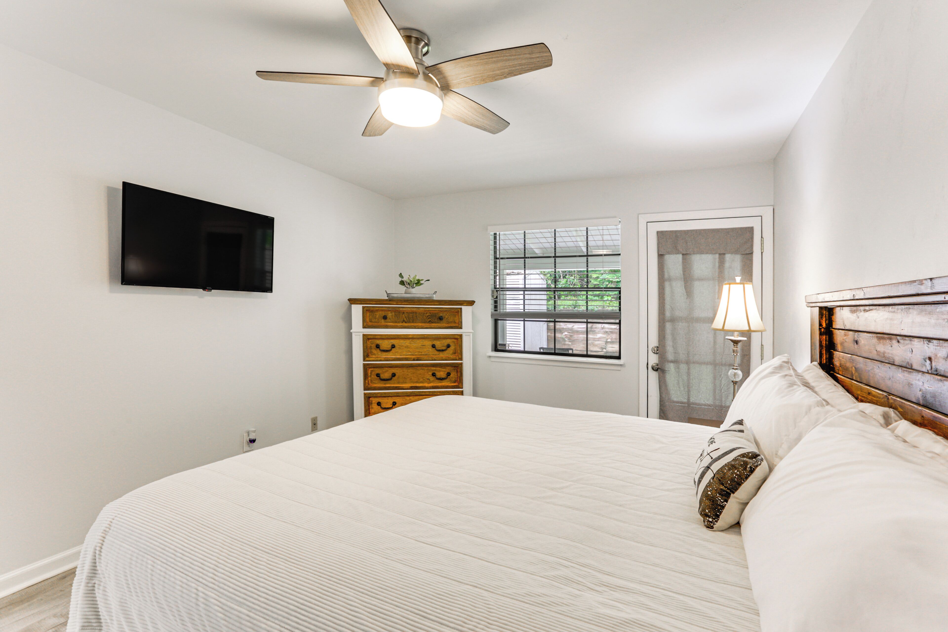 Un dormitorio con cama, ventilador de techo, televisor montado en la pared, cómoda, ventana y puerta.