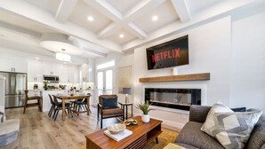 A modern living room with a fireplace, a television mounted above it, and a dining area in the background.