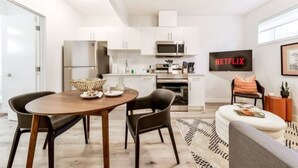 A modern kitchen with white cabinets, a microwave, and a coffee machine. There is a dining area with a wooden table and chairs, and a sofa with a patterned rug. A Netflix sign is visible on the wall.