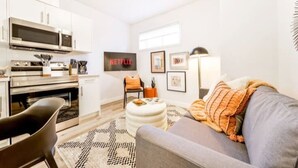 A compact living space with a kitchenette, a grey sofa, a patterned rug, and a wall-mounted TV displaying the Netflix logo.