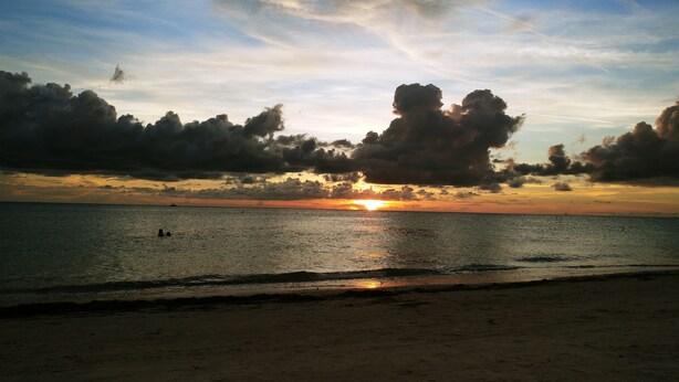 Een strand met een zonsondergang boven de oceaan, wolken aan de hemel en een paar mensen in het water.