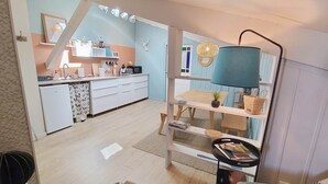 Una cocina moderna con armarios blancos, un microondas y una cafetera. Hay un comedor con una mesa, sillas, una lámpara y una planta pequeña.