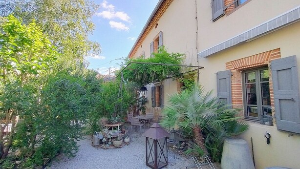 Un patio con una mesa y sillas, rodeado de follaje y un edificio con ventana y persiana.