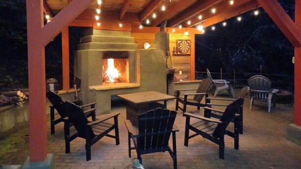 An outdoor patio with a stone fireplace, wooden chairs, and string lights. - Laurel Ridge, Privacy yet minutes from downtown, Dupont & Pisgah National Forest (Pisgah Forest)