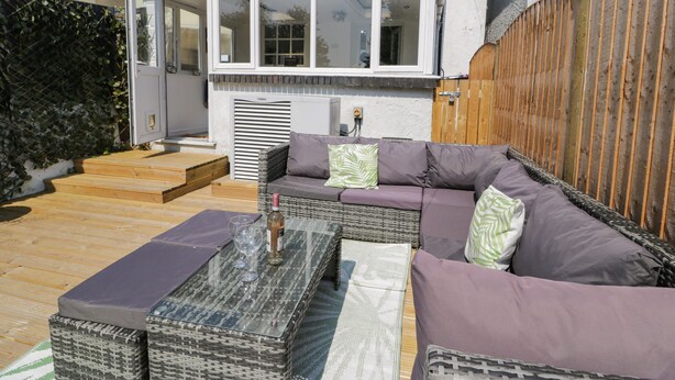 A patio with a wicker sofa, a glass coffee table, and a wooden deck. - Penrallt (Cemaes Bay)