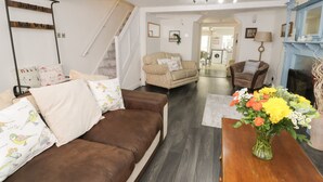 A spacious living room with a brown leather sofa, a beige armchair, and a wooden coffee table with a vase of flowers. A staircase leads to an upper floor, and a kitchen is visible in the background. - Penrallt (Cemaes Bay)