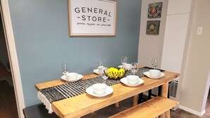 A dining area with a wooden table set for four, white plates, and glasses. A framed sign above reads "GENERAL STORE" with a list of items. - Tranquil 3 Bedroom, 2 Bath Home Near Scenic River Walk! (Baker City)