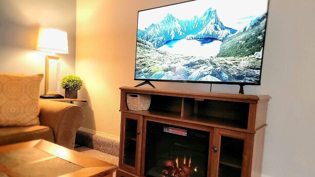 A living room with a flat-screen TV mounted on a wooden stand, a fireplace below, a sofa, a lamp, and a small plant. - Tranquil 3 Bedroom, 2 Bath Home Near Scenic River Walk! (Baker City)