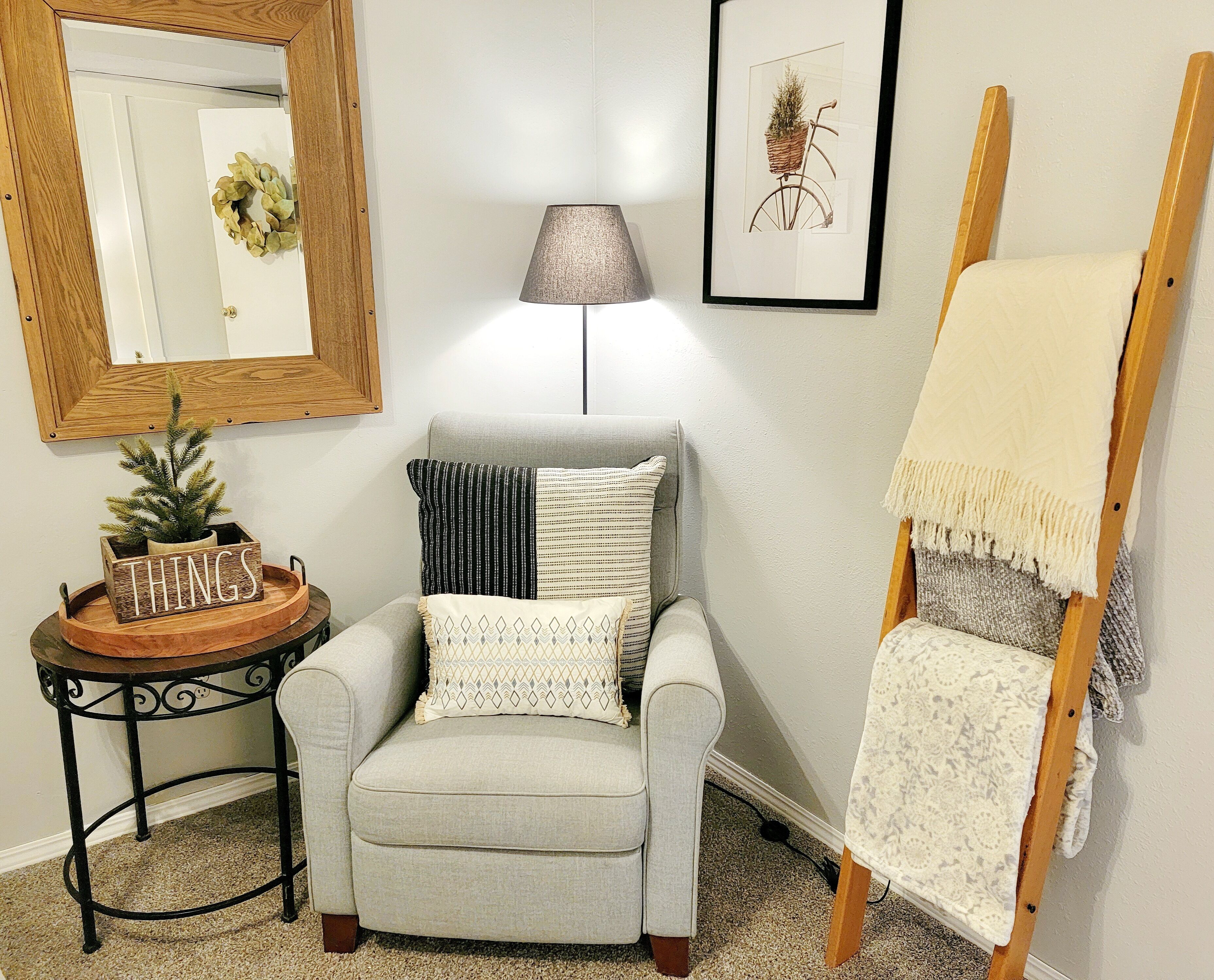 Una habitación con un sillón gris, una pequeña mesa con un cartel que dice 'THINGS', un cuadro con una bicicleta, una lámpara y un caballete con un lienzo en blanco.