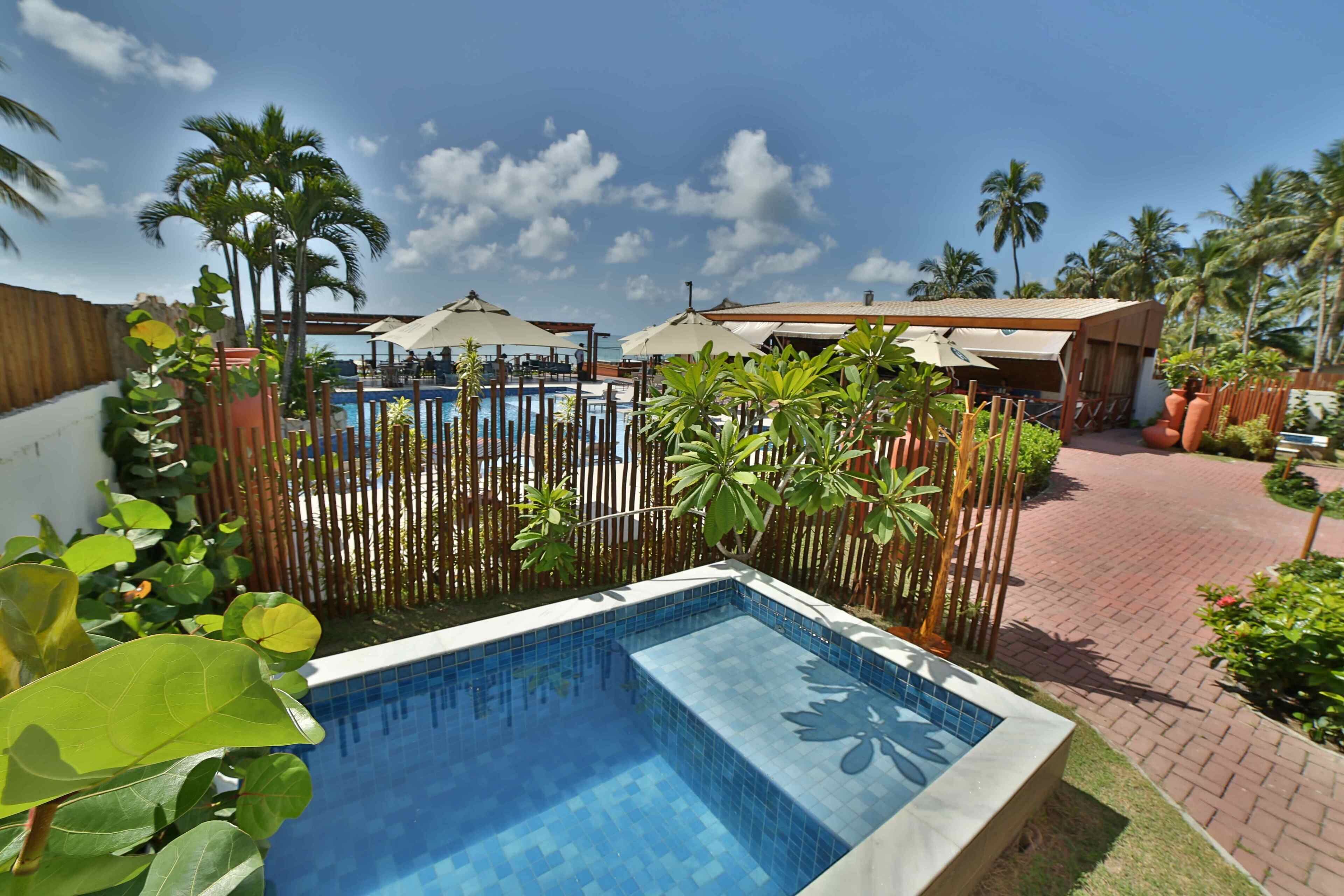 Une piscine entourée de plantes tropicales et délimitée par une clôture en bois, avec un ciel bleu clair en arrière-plan.