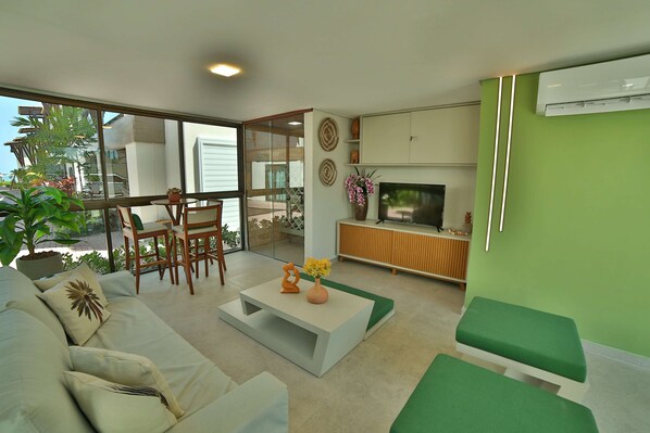 Una sala de estar moderna con sofá, mesa de centro y mueble para televisión. Hay un balcón con mesa y sillas, y un ventanal con vistas al exterior.