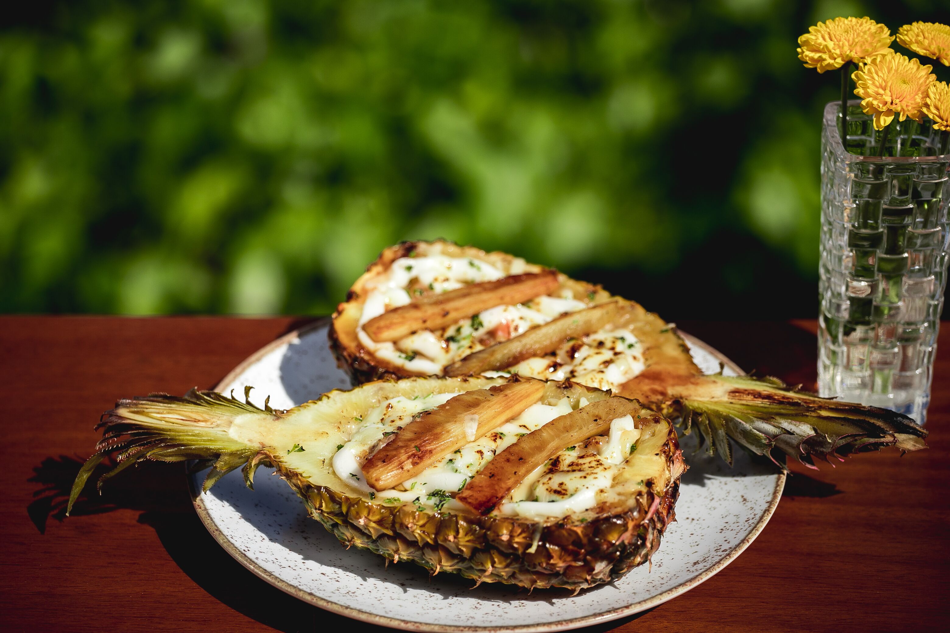 A plate with two grilled pineapple halves topped with a creamy sauce and herbs, accompanied by a glass of water and a vase with yellow flowers.