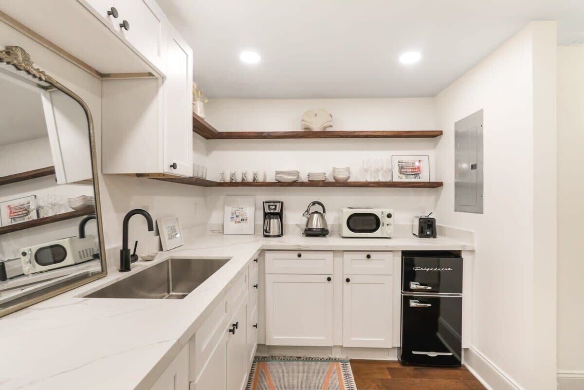 Una cocina moderna con armarios blancos, un lavamanos de acero inoxidable, una cafetera, un hervidor, un microondas y un refrigerador negro.