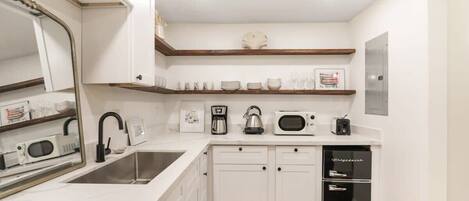 Una cocina moderna con armarios blancos, un lavamanos de acero inoxidable, una cafetera, un hervidor, un microondas y un refrigerador negro.