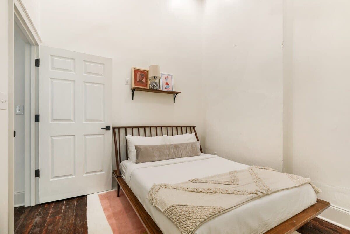 Een slaapkamer met een bed, een deur, een plank met foto’s en een lamp, en een houten vloer.