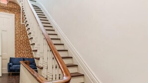 Eine Treppe mit Holzboden und weißen Balustraden führt zu einer Zwischenebene mit einer Ziegelwand und einem blauen Stuhl.