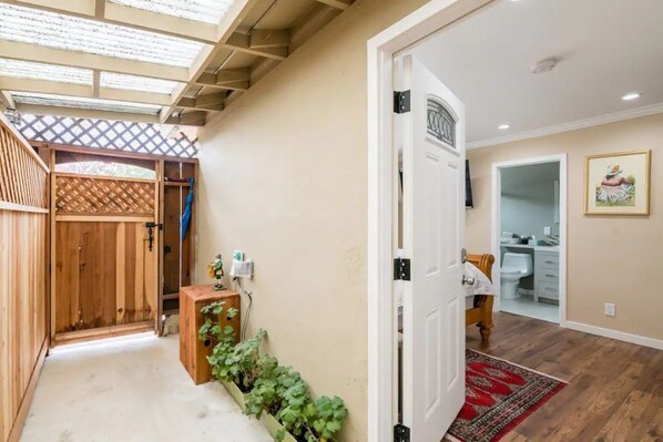 A hallway with a wooden door, a small wooden cabinet with plants, and a red patterned rug leading to a bathroom.