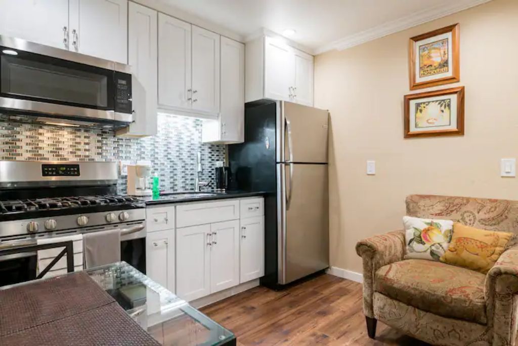 A modern kitchen with white cabinets, a stainless steel stove, and a black refrigerator. There is a glass table and a patterned armchair.