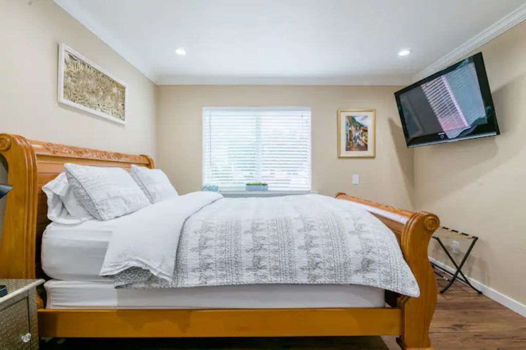 A bedroom with a wooden bed, a flat-screen TV mounted on the wall, and a window with blinds.