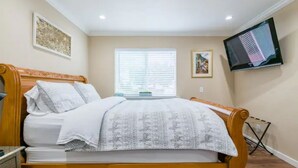 A bedroom with a wooden bed, a flat-screen TV mounted on the wall, and a window with blinds.