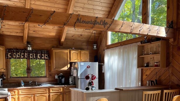 Cocina de cabaña de madera con un ventanal grande, armarios de madera, un refrigerador y una zona de comedor con mesa y sillas.