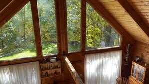 Una cabaña con ventanas amplias que ofrecen vista a un bosque. En su interior, hay una mesa de comedor de madera con sillas, un área de cocina con un hervidor y un armario con puertas de vidrio.