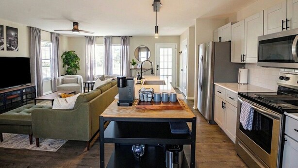 A modern kitchen with stainless steel appliances, a dining area with a wooden table, and a living room with a sofa and a TV. - Few Mins to Ballantyne-Pet Friendly-1 Level Home (Pineville)