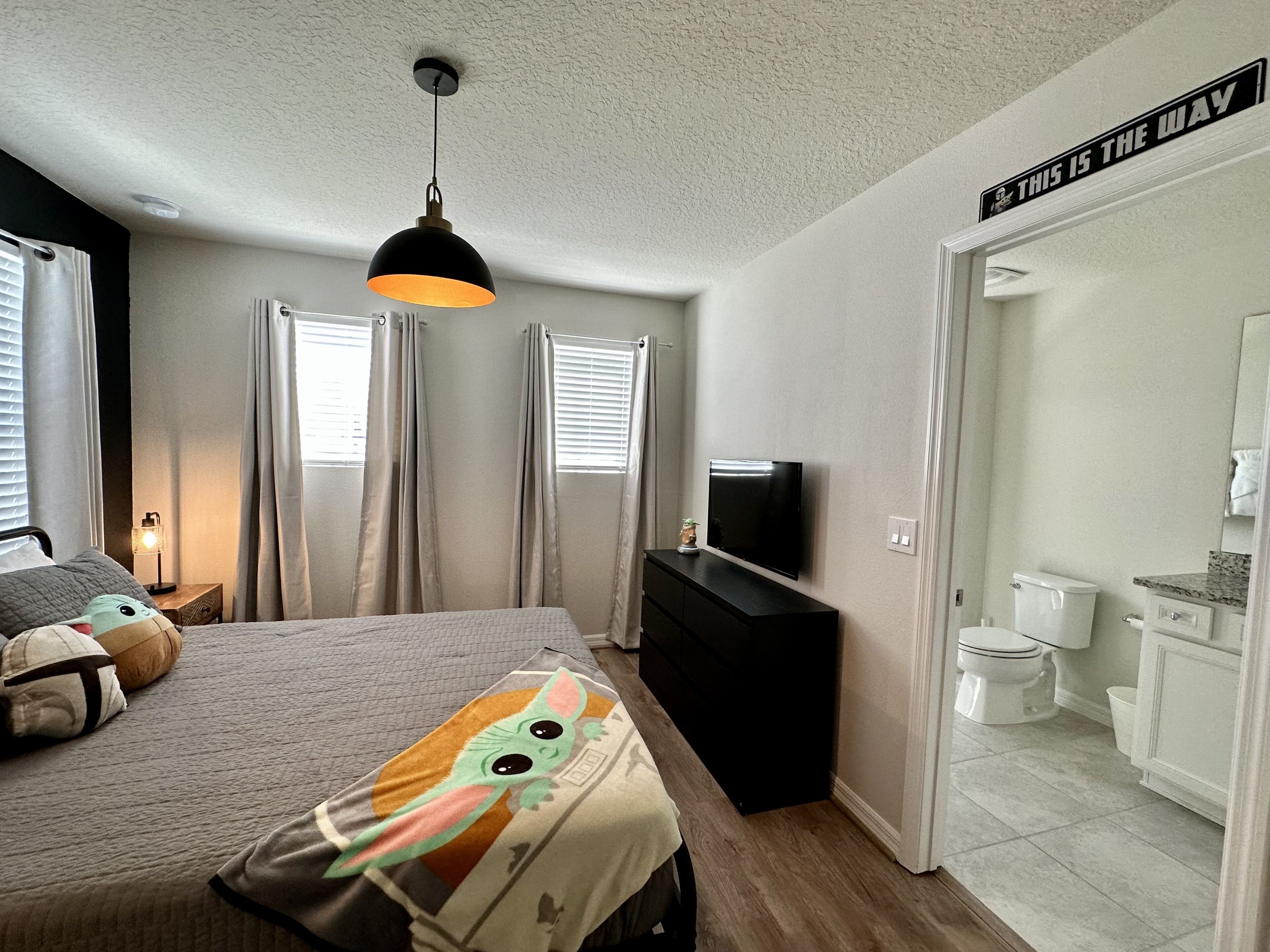 Une chambre à coucher avec un lit, un téléviseur posé sur une commode noire, une salle de bain avec des toilettes et un lavabo, et une pancarte sur la porte indiquant : « C’EST LA BONNE DIRECTION ».