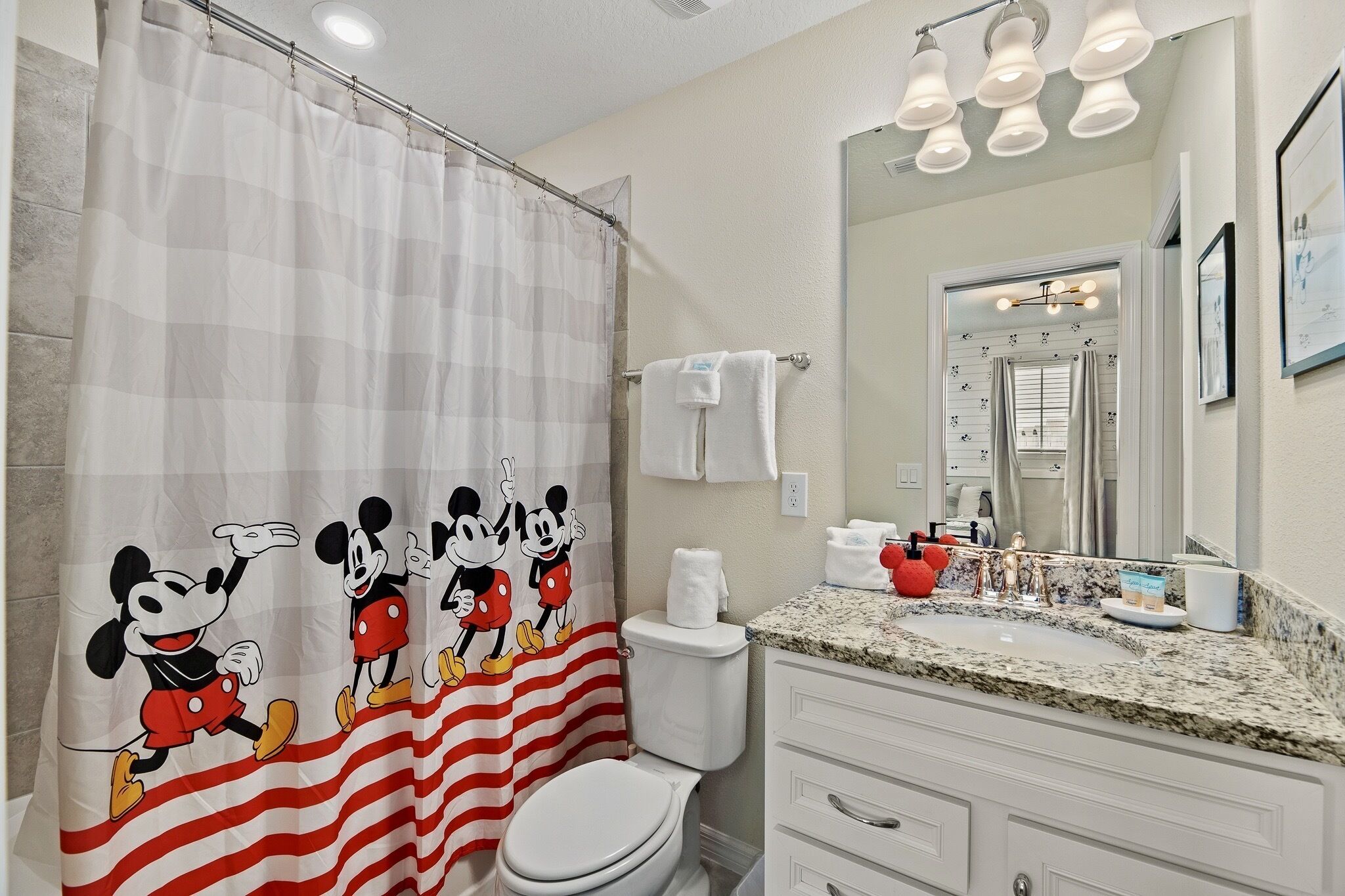 Un baño con cortina de ducha de Mickey Mouse, un inodoro, un lavamanos con encimera de granito y un espejo sobre el lavamanos.