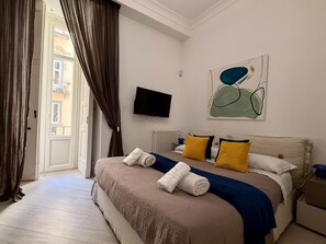 A hotel room with a bed, a television, a painting, and a view through a door.