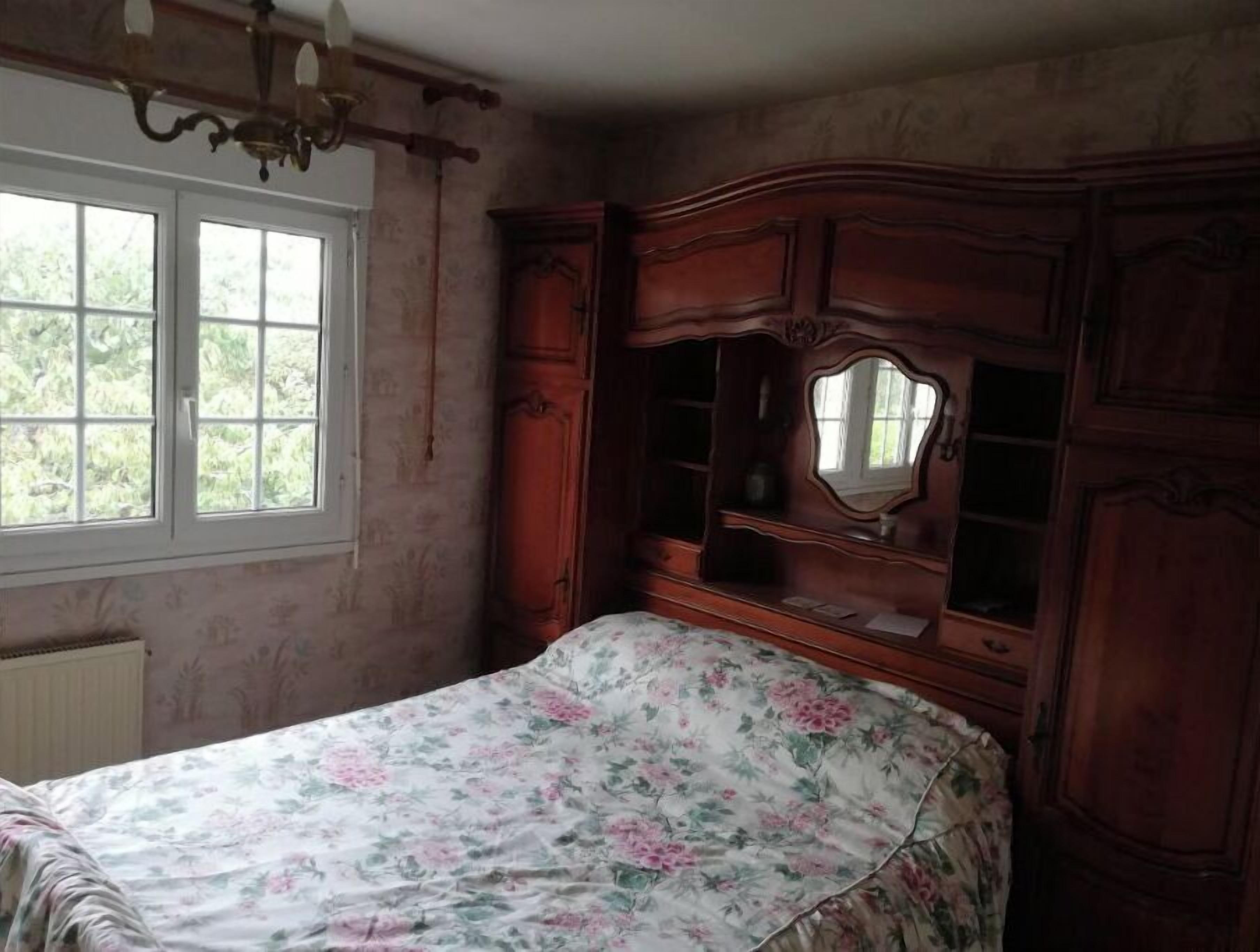 A bedroom with a floral bedspread, a wooden dresser with a mirror, and a window with a view of greenery.