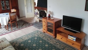 A living room with a television on a wooden stand, a fireplace, a dining table with chairs, and a patterned rug.