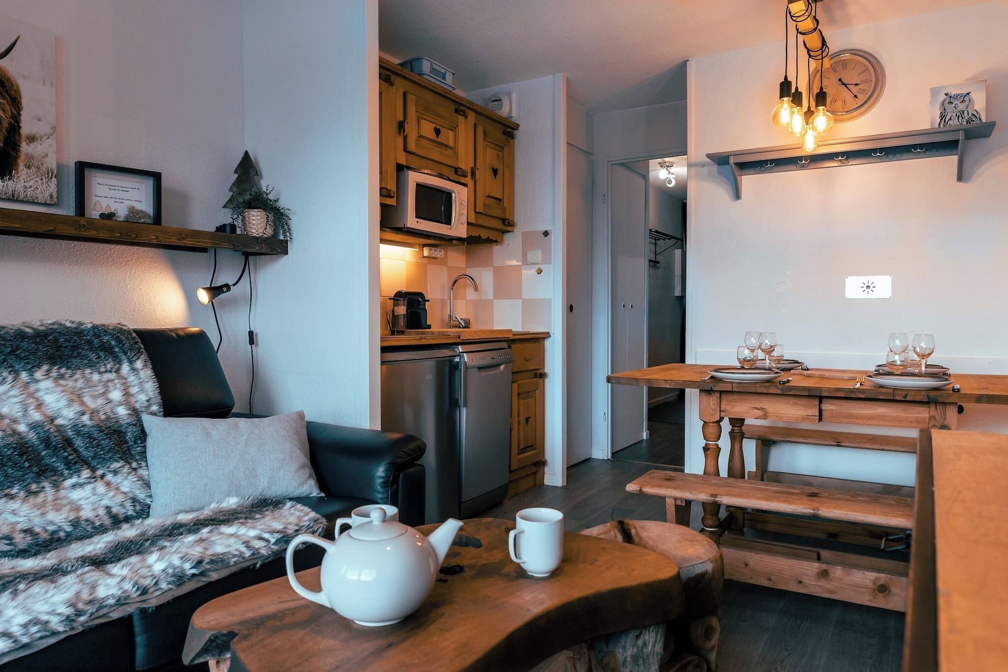 A cozy living room with a sofa, a coffee table, and a wooden bench. A kitchen area with a microwave, a sink, and a refrigerator is visible in the background.