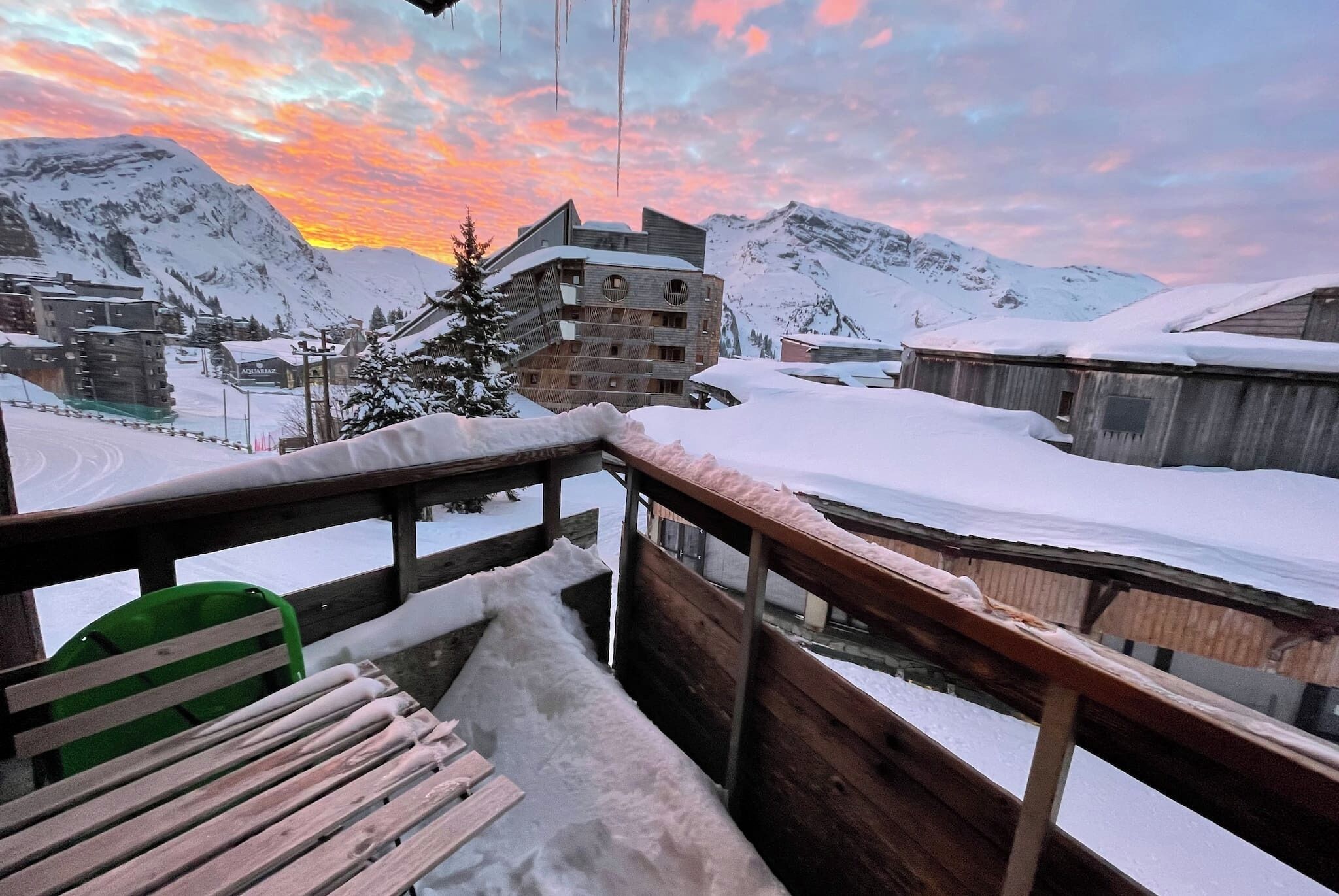 A wooden deck with snow-covered ground, overlooking a mountainous area with buildings and a sunset sky.