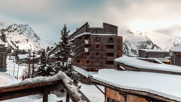 Un edificio a più piani con facciata in legno e tetto spiovente, circondato da montagne innevate e un cielo nuvoloso.