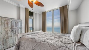 A bedroom with a large bed, a ceiling fan, and a view of the sea through large windows.