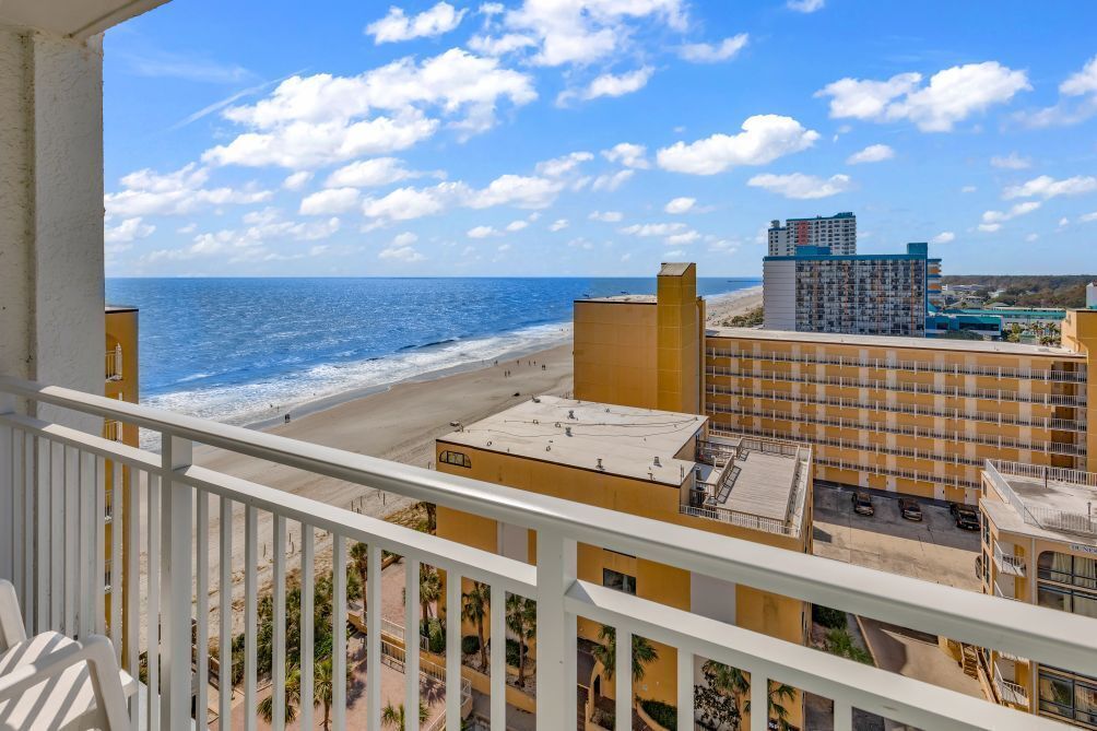 A balcony with a view of a beach and ocean, buildings in the background, and a clear sky.
