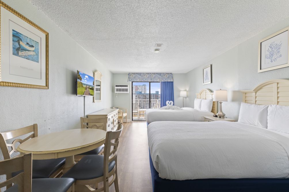 A hotel room with two beds, a dining area with a table and chairs, a TV, and a balcony with a view of the city.