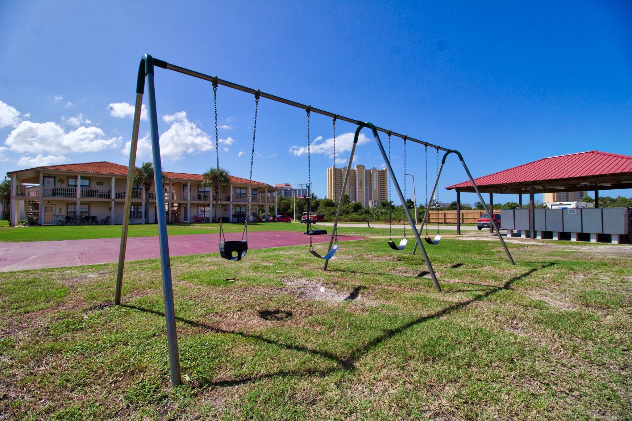 Un parc infantil amb cadira de balanç, un edifici amb teulada vermella i un complex residencial de diversos pisos al fons.