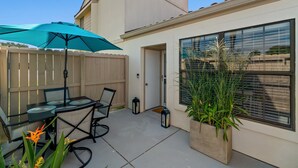 Une terrasse avec une table et des chaises, un parasol de couleur turquoise et un jardinier avec des plantes.