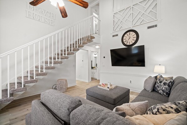 A modern living room with a staircase, grey sofas, a flat-screen TV, and a clock on the wall.