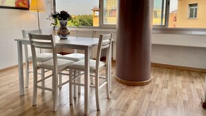 Un comedor con una mesa y sillas blancas, un florero con flores y un ventanal con vistas a edificios.