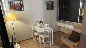 Una habitación con una mesa de comedor, dos sillas, una mesita auxiliar con una lámpara, un ventanal grande y dos cuadros en la pared.