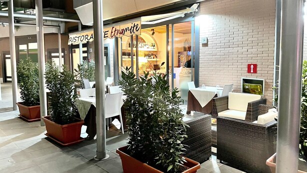 Un área de estar al aire libre con plantas en macetas, mobiliario de mimbre y la entrada de un restaurante con un letrero.