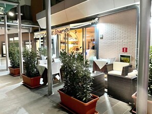 An outdoor seating area with tables and chairs, potted plants, and a restaurant with a brick wall and glass doors.